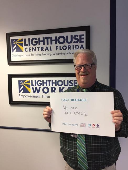 An image of Todd at the Lighthouse Works office holding a sign that says 'We are all one'