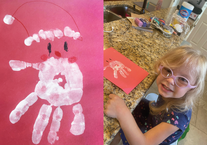 Maddalena smiling for the camera in her family's kitchen with her artwork depicting a Santa Claus on red construction paper with a palm print in white paint as his face and beard