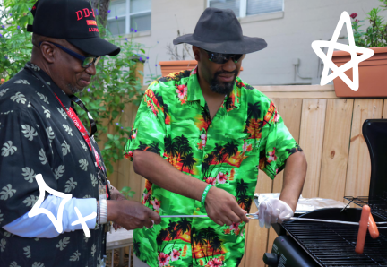 250 Two men stand together by a grill cooking hot dogs. One is wearing gloves and a hat while holding tongs.