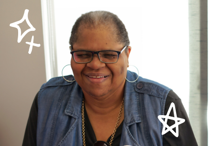 50 A woman wearing glasses and hoop earrings smiles warmly while sitting indoors.