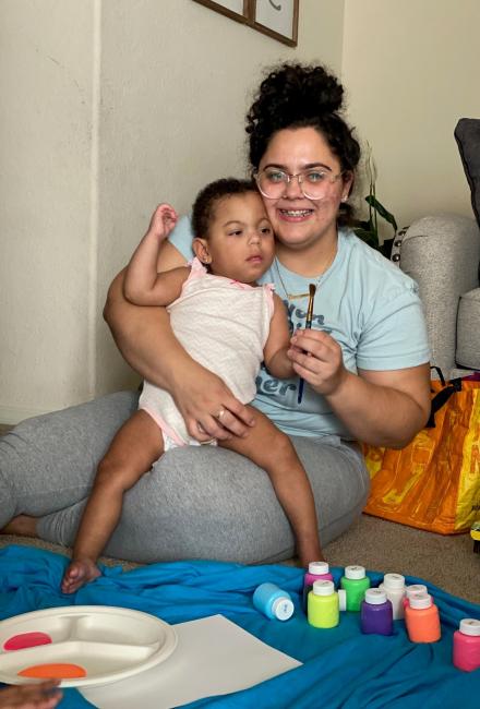 Marlie sitting on Mom's lap, holding a paint brush before starting their art activity