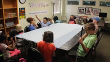 A group of elementary School age children gathered around a table raising their hands to answer a question in our School Age Children services program