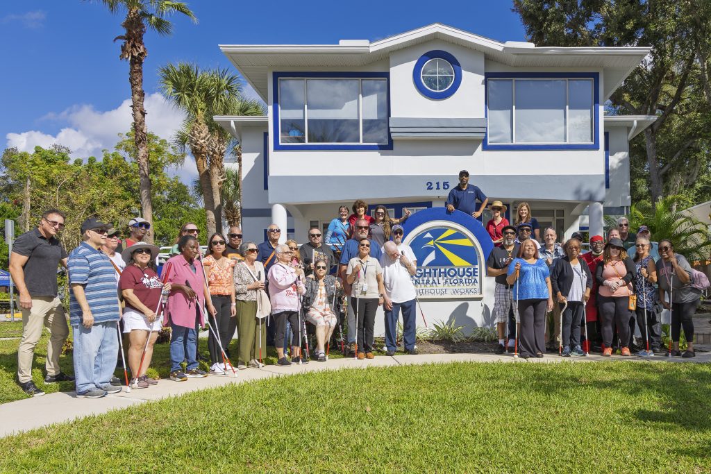 A group of people standing together outside of Lighthouse Central Florida. They are all smiling and standing close together. Some are wearing sunglasses and carrying white canes.