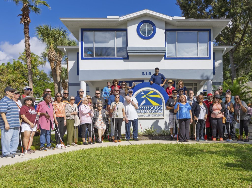A group of people standing together outside of Lighthouse Central Florida. They are all smiling and standing close together. Some are wearing sunglasses and carrying white canes.
