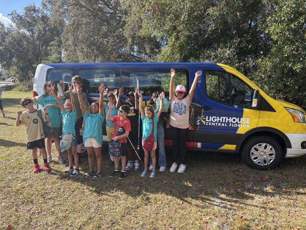 Kids vans Group of children standing and waving beside a blue-and-yellow Lighthouse Central Florida van in a grassy park area.