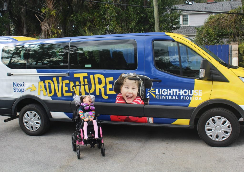 Sailor A smiling child in a wheelchair sits in front of a blue-and-yellow Lighthouse Central Florida van featuring large “Next Stop Adventure” text and a photo graphic.