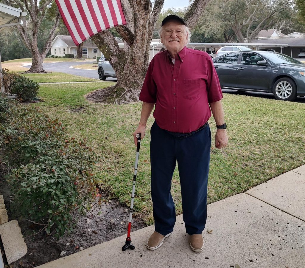 Older man wearing a red shirt and cap stands on a sidewalk holding a cane, smiling, with an American flag hanging from a tree in a residential neighborhood.
