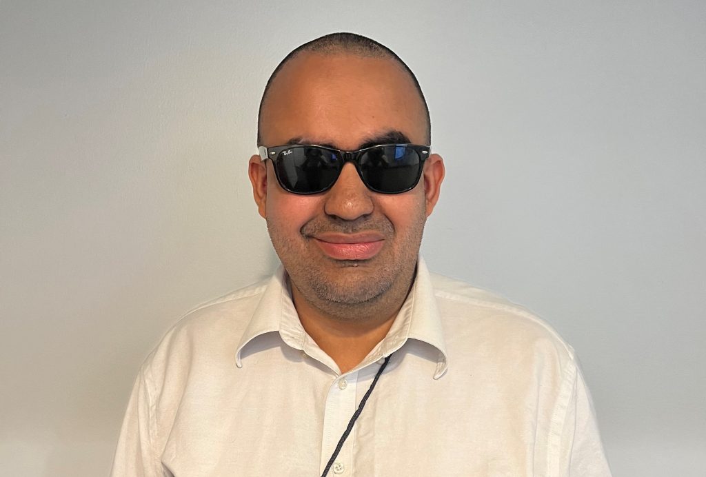 Man wearing dark sunglasses and a white collared shirt, smiling slightly while facing the camera against a plain light-colored wall.