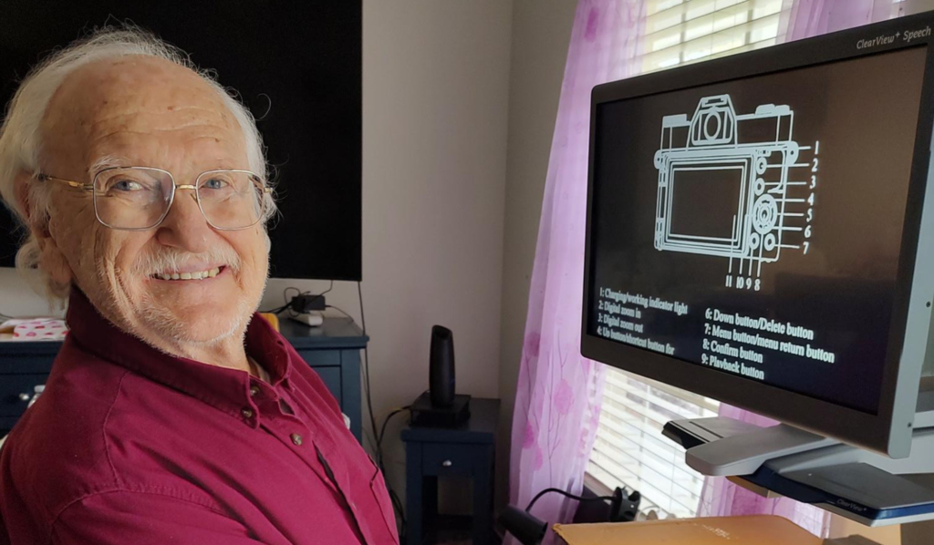 Older man with glasses smiles at the camera while seated at a desk, next to a monitor showing a labeled diagram of a digital camera.