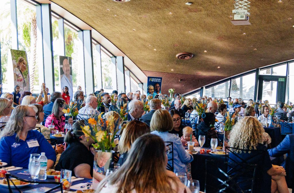 A large group of people sit inside a room with tall windows at large tables filled with flower arrangements, pictures, and food.