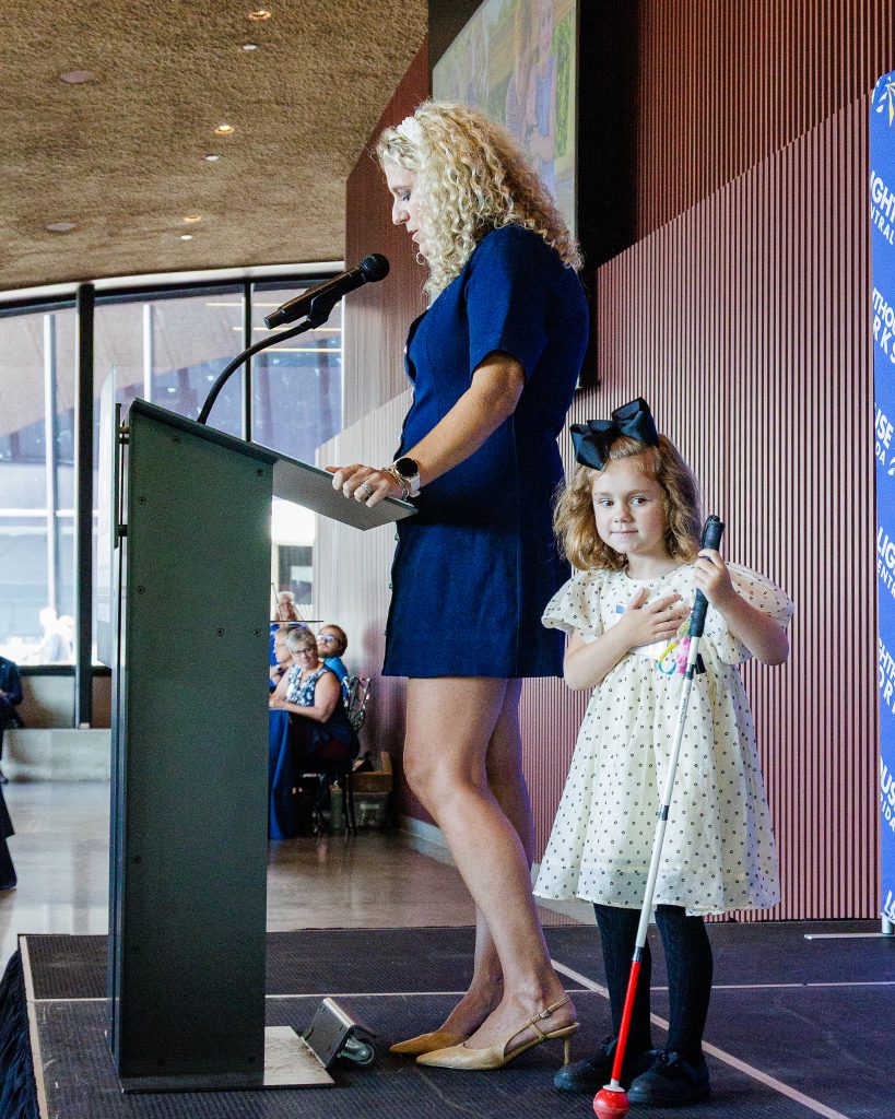 A woman in a blue dress stands on a stage behind a podium and microphone next to a young child in a dress and hairbow holding a white cane.