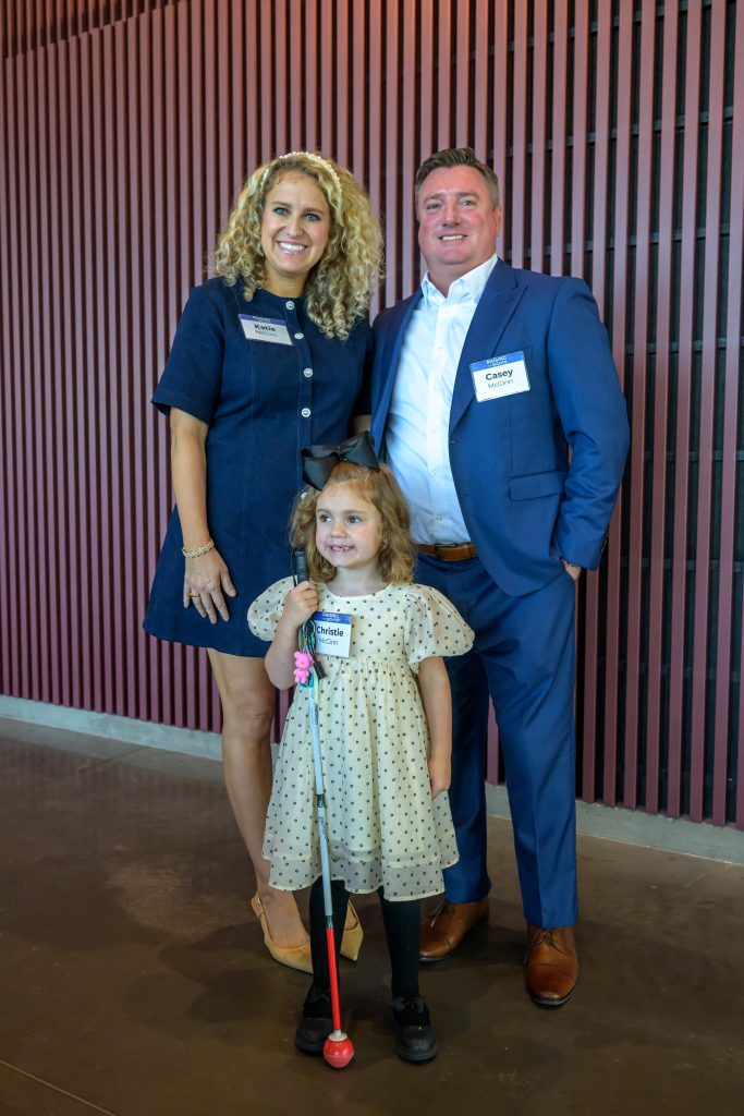 A woman in a blue dress stands smiling next to a man in a blue suit with a young child in a dress and hairbow holding a white cane.