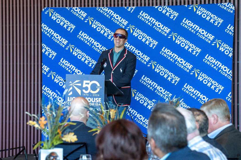 A man in sunglasses and a blazer stands smiling behind a podium and microphone in front of a Lighthouse backdrop.